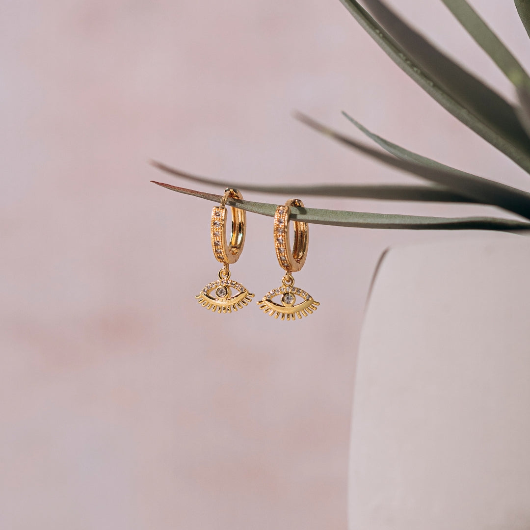 gold evil eye hoop earrings on plant leaf with pink background